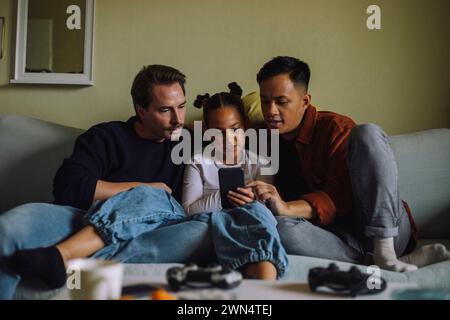 Girl using smart phone while sitting with gay parents on sofa at home Stock Photo