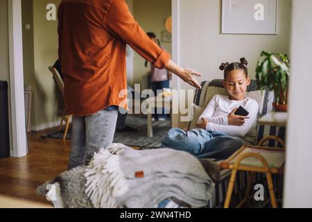 Midsection of father asking smart phone from daughter sitting on bed at home Stock Photo