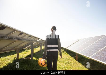 Rear view of female engineer holding hard hat while walking amidst solar panels at power station Stock Photo