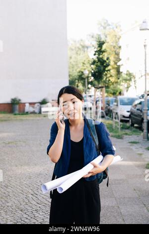 Mature female architect talking on mobile phone while walking with blueprints on footpath Stock Photo