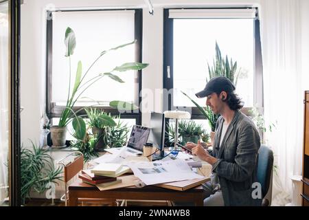 Side view of male architect using smart phone while sitting at desk in home office Stock Photo