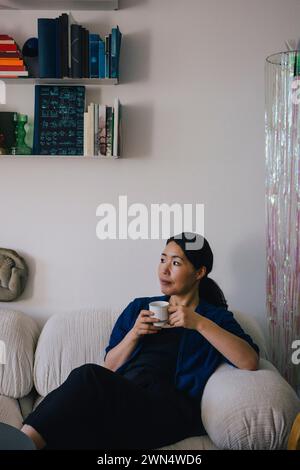 Thoughtful woman with coffee cup sitting on sofa at home Stock Photo