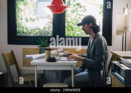 Side view of male architect using laptop at desk in home office Stock Photo