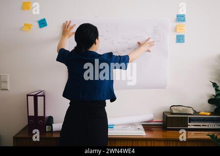Rear view of female architect examining blueprint on wall at home office Stock Photo