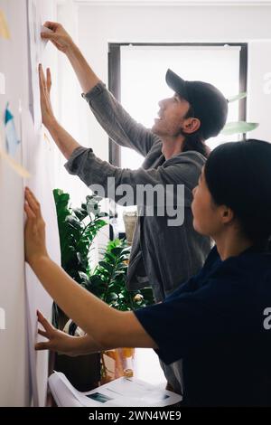 Male and female architect colleagues discussing over blueprint on wall at home office Stock Photo