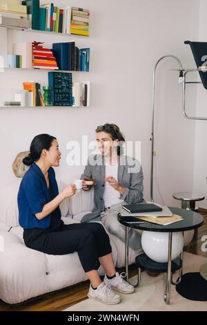 Full length of happy male and female architects with coffee cups sitting on sofa at home office Stock Photo