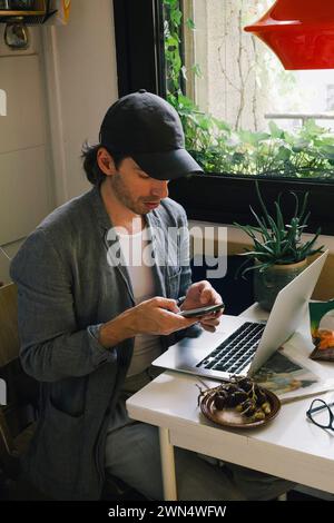 Male architect using smart phone while sitting with laptop at desk in home office Stock Photo