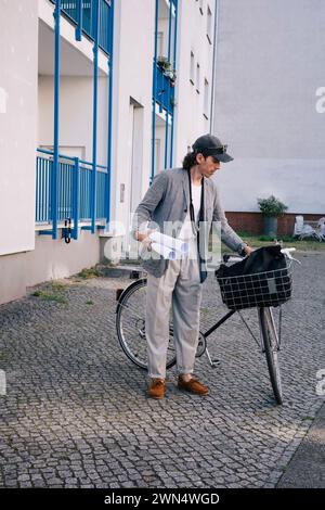 Full length of male architect holding blueprint rolls standing by bicycle on footpath Stock Photo