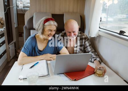 Happy senior couple sitting with laptop at table in camper van Stock Photo