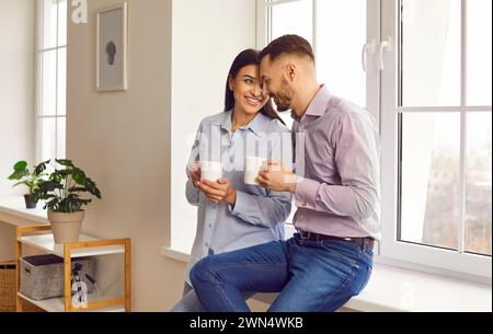 Happy smiling couple sitting on the windowsill at home with a cup of tea or coffee. Stock Photo