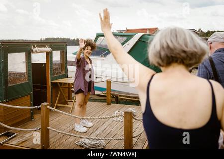 Happy young woman standing on houseboat waving at senior couple Stock Photo