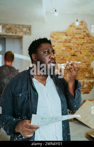Male construction manager with floor plan talking on mobile phone speaker at site Stock Photo