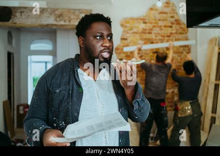 Male construction manager with floor plan talking on mobile phone speaker while workers working in background at site Stock Photo