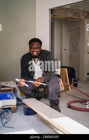 Portrait of cheerful carpenter with measuring instrument and plank in apartment Stock Photo