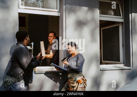 Workers passing plank through window while female construction manager talking on phone outside house Stock Photo