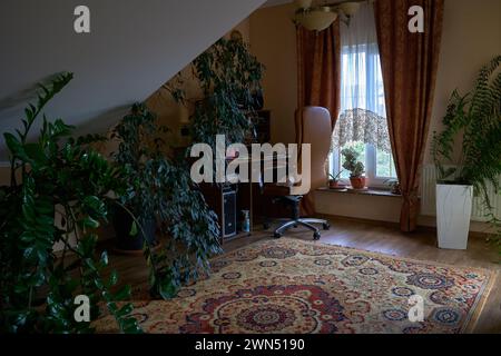 Home workplace: table, computer, green house plants, brown armchair near window. Modern home cozy working place. Real interior. Stock Photo