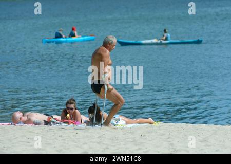 Old one-legged man walking with double adjustable elbow crutches, people sunbathing on a river sandy beach, canoes floating. August 2, 2021. Kyiv, Ukr Stock Photo