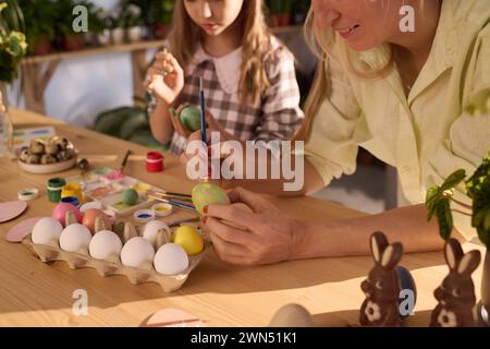 Selective focus shot of mother and daughter sitting at wooden desk in workshop coloring eggs for Easter Stock Photo