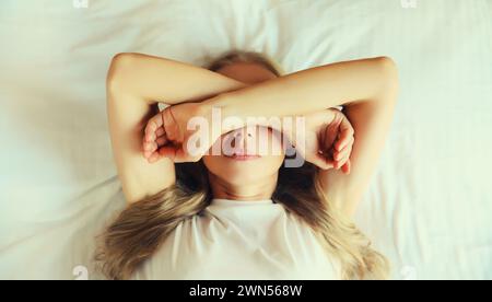 Tired sleepy woman wakes up covering her eyes with her hands lying on the bed on pillow in bedroom at home in the morning Stock Photo