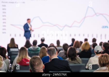 A professional presenter addresses an attentive audience at a business conference with focus on data charts. Stock Photo