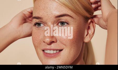 Close up highly detailed portrait of a young smiling woman with freckles. Cropped image of a beautiful blond female adjusting hair against a beige bac Stock Photo