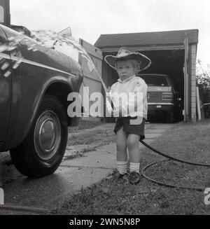 1970s, historical, outside on a grassed driveway, a little girl, wearing a Sheriff hat, holding a garden hose, helping to wash her dad's car, England, UK. A Cortina motorcar of the era parked in a garage further up the incline. Stock Photo