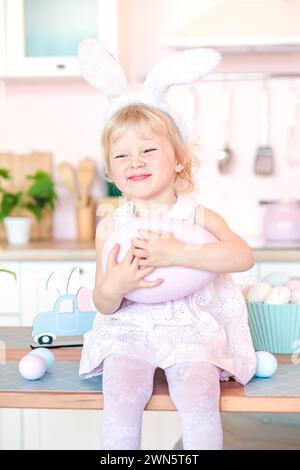 Little funny girl wearing in the ears of an easter bunny in a white dress sitting on a table in the kitchen and holding a huge pink painted easter egg Stock Photo