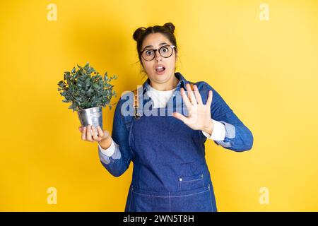 Young caucasian gardener woman holding a plant isolated on yellow background afraid and terrified with fear expression stop gesture with hands, shouti Stock Photo