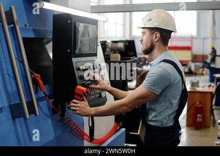 Side view of young bearded Caucasian man wearing hardhat operating modern CNC machine Stock Photo