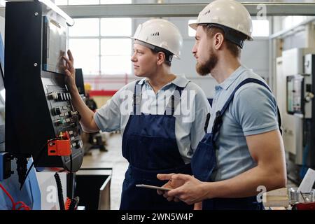 Side view medium shot of male and female engineers wearing hardhats working in factory using CNC machine Stock Photo