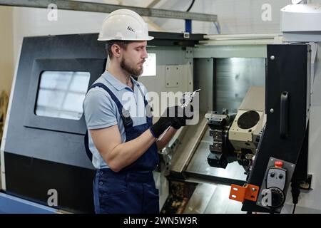 Medium shot of young bearded turner standing in workshop checking size of metal part, copy space Stock Photo