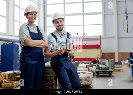 Medium long portrait of successful Caucasian male and female engineers wearing uniform posing for camera in factory workshop Stock Photo
