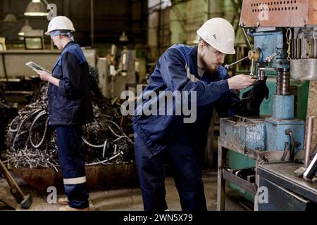 Man and woman wearing uniform working in industrial workshop using digital tablet and old-fashioned equipment Stock Photo