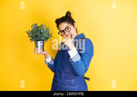 Young caucasian gardener woman holding a plant isolated on yellow background Punching fist to fight, aggressive and angry attack, threat and violence Stock Photo
