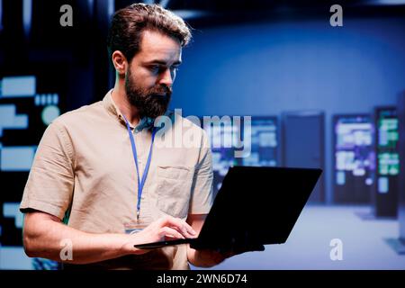 IT admin in hardware room containing server rackmounts, storage devices and networking units providing vast amounts of computing resources. Supervisor doing software maintenance using laptop Stock Photo