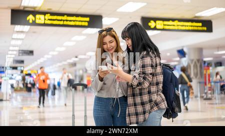 Two cheerful Asian female friends are talking and checking their flight information on their smartphone while standing in the airport. Stock Photo