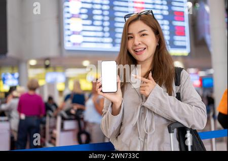 An attractive Asian woman showing a white-screen smartphone mockup to the camera while standing in the airport. people and transportation concepts Stock Photo
