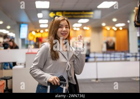 A beautiful Asian female passenger is waving her hand to say goodbye to her friends in the airport, traveling by plane. Stock Photo