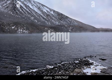 A view from a rocky shore to a large lake at the foot of a mountain range over which evaporation rises on a cloudy autumn day. Lake Kok-Kol, Altai, Si Stock Photo