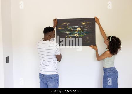 Biracial African American couple hang a painting on a white wall at home Stock Photo