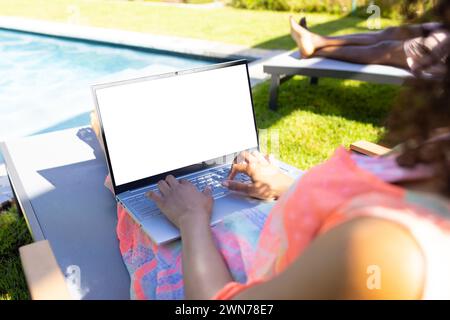 Young biracial woman works on a laptop with a blank screen by the poolside at home, with copy space Stock Photo