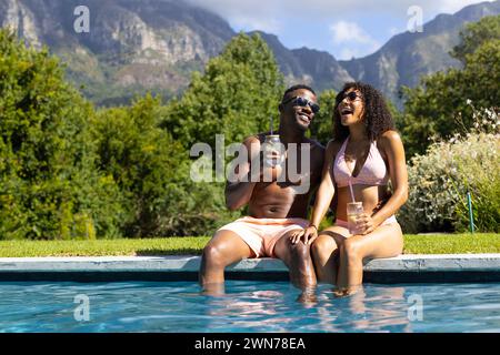 African American man and biracial woman enjoy drinks by the pool, surrounded by mountains Stock Photo