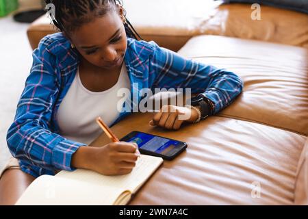 Young African American woman writes in notebook using her smartphone for booking vacation online Stock Photo