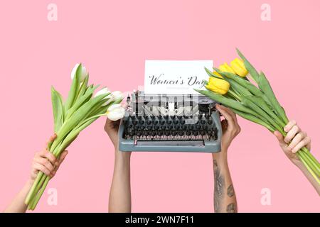 Hands holding vintage typewriter, tulips and festive postcard with text WOMEN'S DAY INTERNATIONAL on pink background Stock Photo