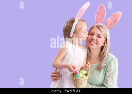 Little girl with Easter eggs kissing her mother on lilac background Stock Photo