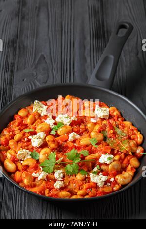 gigantes plaki, greek giant baked beans in a chunky tomato sauce sprinkled with feta cheese and fresh cilantro on skillet on black wooden table, verti Stock Photo