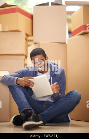Boxes, home and black man with a tablet, smile and typing with internet and package in lounge. African person on the floor, apartment and guy with Stock Photo