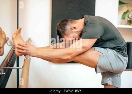 Side view of barefoot muscular male looking down while standing in gym stretching leg with hands together on barre near mirror in daylight Stock Photo