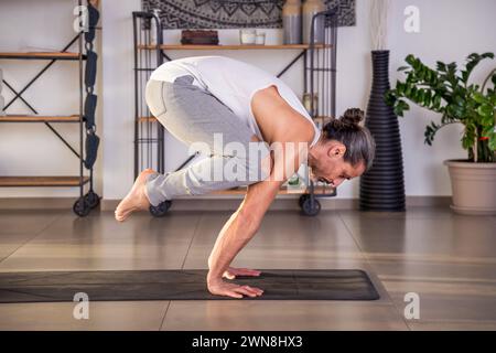 Side view of fit guy with ponytail practicing Kakasana pose on mat during yoga session in spacious studio with shelves and potted green plant on backg Stock Photo