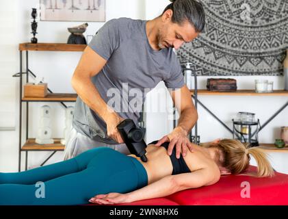 Experienced fitness trainer using massage gun on lower back of woman lying on stretcher during recovery process in modern studio Stock Photo